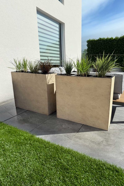 Zwei rechteckige Pflanzgefäße mit horizontaler Rillenstruktur auf einer Terrasse, bepflanzt mit Gräsern vor einer Hauswand.