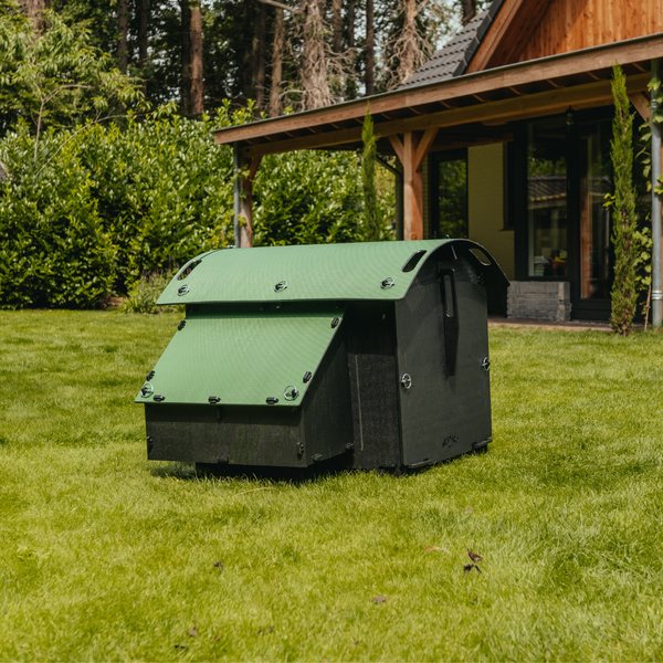 Ein grün-schwarzer Hühnerstall aus Kunststoff steht auf einer Rasenfläche in einem Garten vor einem Holzhaus.