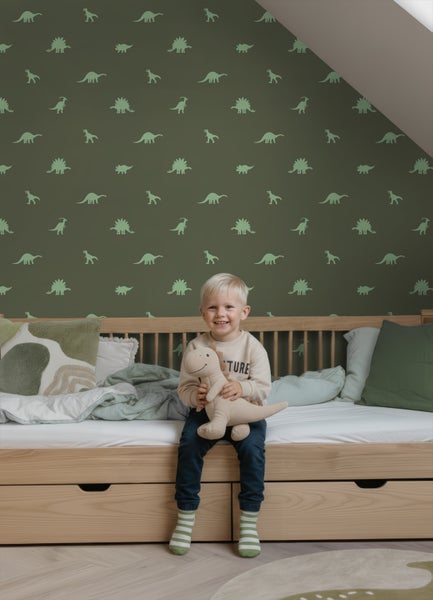 Helles Kinderzimmer mit dunkelgrüner Dinosaurier-Tapete, einem hellen Holzbett mit Schubladen und einem Kind mit einem Dinosaurier-Stofftier.