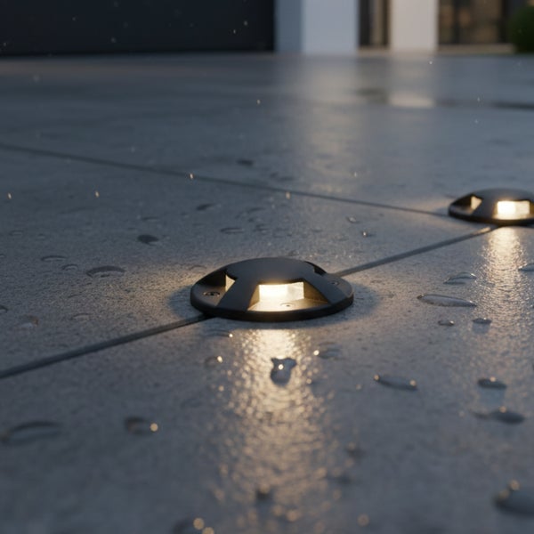 Zwei runde, schwarze LED-Bodenstrahler auf grauen, nassen Terrassenplatten bei Dämmerung mit warmweißem Lichtschein.