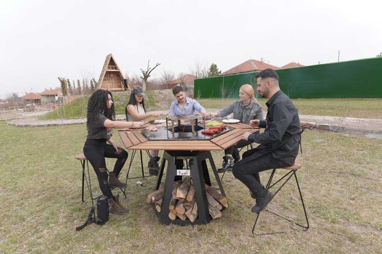 Eine Gruppe von Freunden bereitet gemeinsam Grillgut auf einem achteckigen Grilltisch mit integrierter Feuerstelle in einem Garten zu.