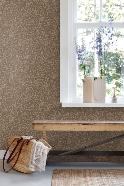 Helle, rustikale Raumszene mit floral gemusterter Tapete, einer Holzbank und einem Fenster mit Blumen in strukturierten Vasen.