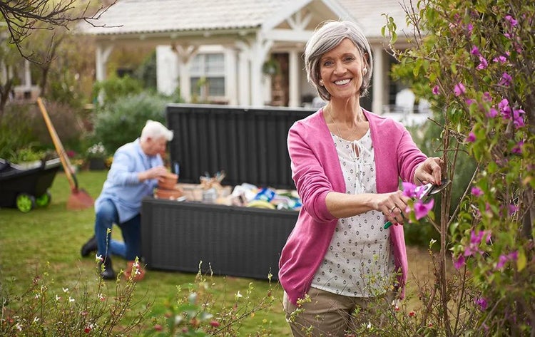 Frau beschneidet einen Strauch mit einer Gartenschere, während im Hintergrund ein Mann eine große schwarze Gartenbox in einem Garten einräumt.
