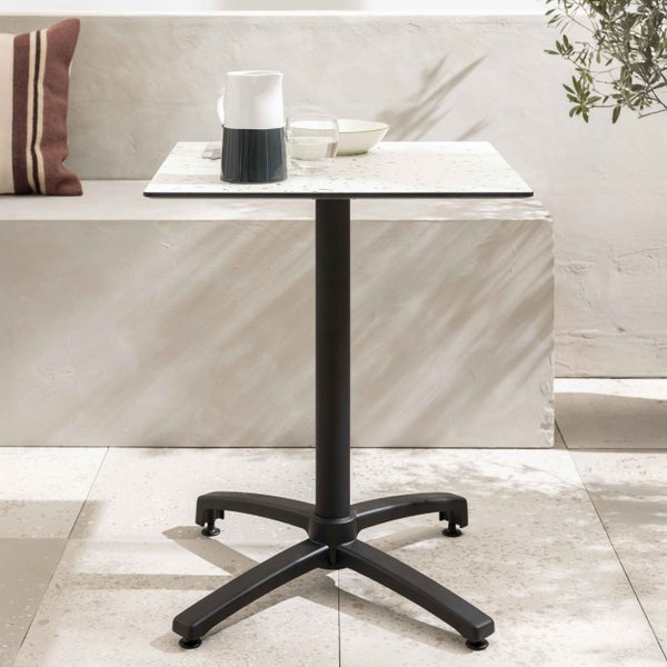 Quadratischer Bistrotisch mit weiß gesprenkelter Tischplatte und schwarzem Metallgestell auf einer Terrasse mit Geschirr.