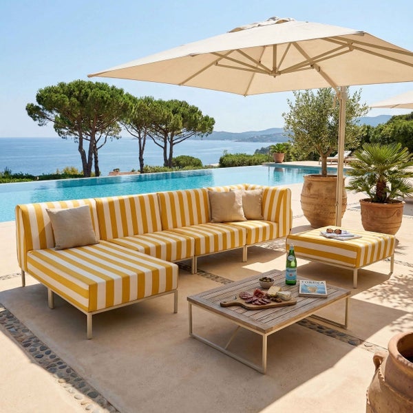 Moderne Gartenlounge mit gelb-weiß gestreiftem Sofa, Hocker und Holztisch an einem Pool mit Meerblick unter einem Sonnenschirm.