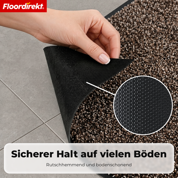 Floordirekt Logo. Eine Hand hebt die Ecke einer Fußmatte an, um die strukturierte, rutschhemmende Unterseite auf Fliesen zu zeigen. Detailansicht.