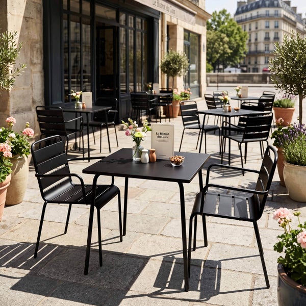 Außenbereich eines Bistros mit quadratischen Metalltischen und Metallstühlen auf einer Steinterrasse, umgeben von Pflanzgefäßen mit Blumen.