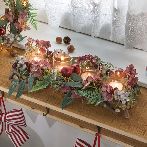 Weihnachtliches Tischgesteck mit vier Teelichtern in Glasbechern dekoriert mit Kunstblumen, Zweigen, Zapfen und Weihnachtskugeln auf Holzbasis.