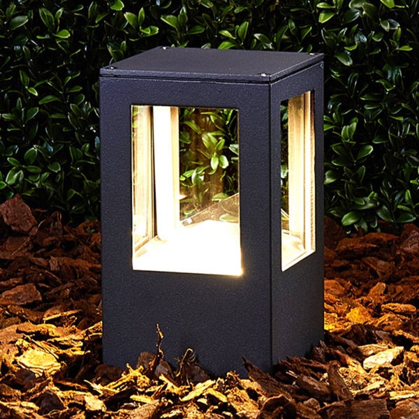 Moderne LED-Sockelleuchte in Anthrazit mit warmweißem Licht, platziert in Rindenmulch vor einer grünen Hecke im Garten.