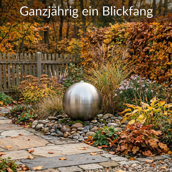 Garten mit Edelstahl-Kugelbrunnen auf Flusssteinen, umgeben von herbstlichen Pflanzen, Gräsern, einem Holzzaun und Steinweg.