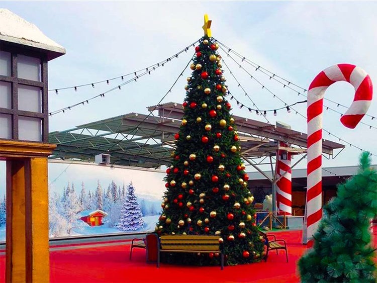 Weihnachtsdekoration im Außenbereich mit geschmücktem Tannenbaum, Lichterkette, Sitzbank und großer Zuckerstangenfigur auf rotem Untergrund.