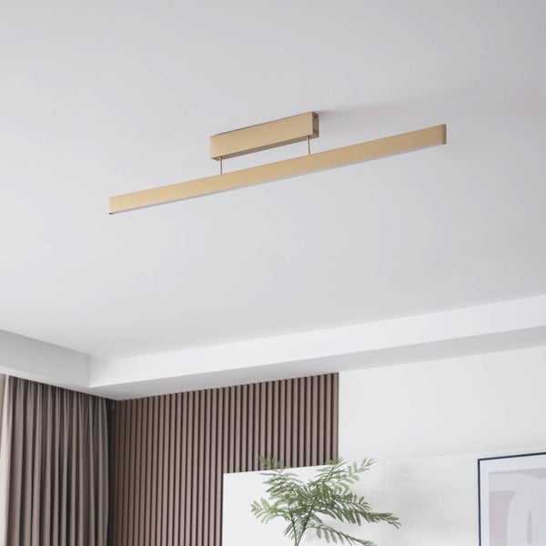 Moderne lineare LED-Deckenleuchte in Goldoptik, montiert an einer weißen Zimmerdecke in einem hellen Raum mit Lamellenwand und Pflanze.