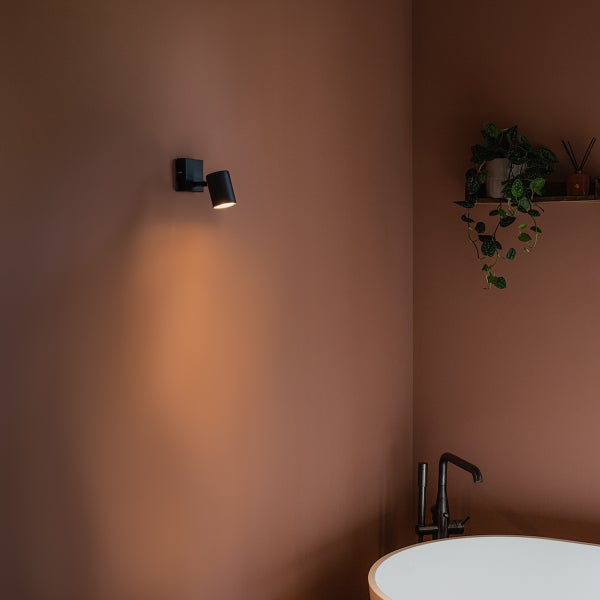 Badezimmer mit Wandleuchte, Badewanne und Pflanze auf einem Regal