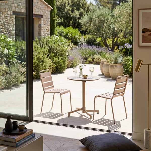 Modernes Garten-Set aus Metall mit zwei Lattenstühlen und einem runden Bistrotisch auf einer sonnigen, mediterranen Terrasse mit üppiger Bepflanzung.