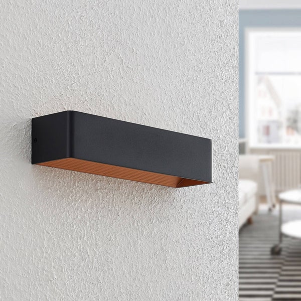 Moderne rechteckige Wandleuchte an einer strukturierten Wand in einem hellen Raum. Schwarze Außenseite und kupferfarbene Innenseite.
