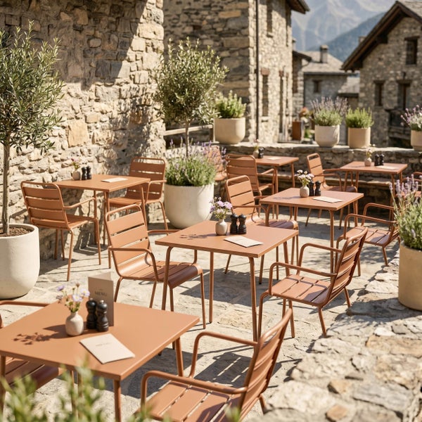 Terrasse mit terracottafarbenen Metalltischen und Gartenstühlen auf Steinboden, umgeben von Natursteinmauern und Pflanzenkübeln.