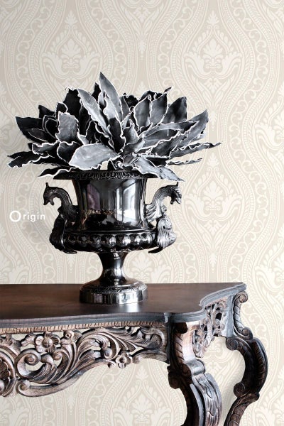 Dekorative Pflanze in einem verzierten silberfarbenen Topf auf einem verzierten Holztisch vor einer gemusterten Tapete.