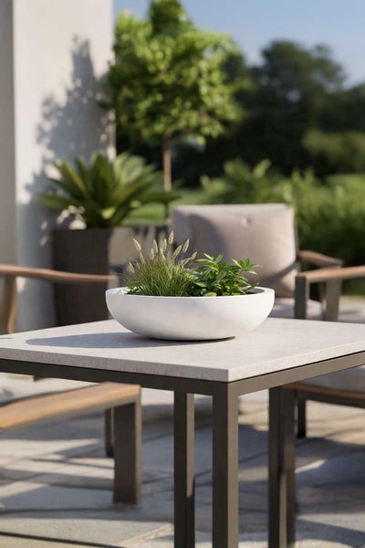 Flache weiße Pflanzschale mit grünen Pflanzen auf einem modernen Gartentisch aus Stein und Metall in einer hellen Terrassenumgebung.