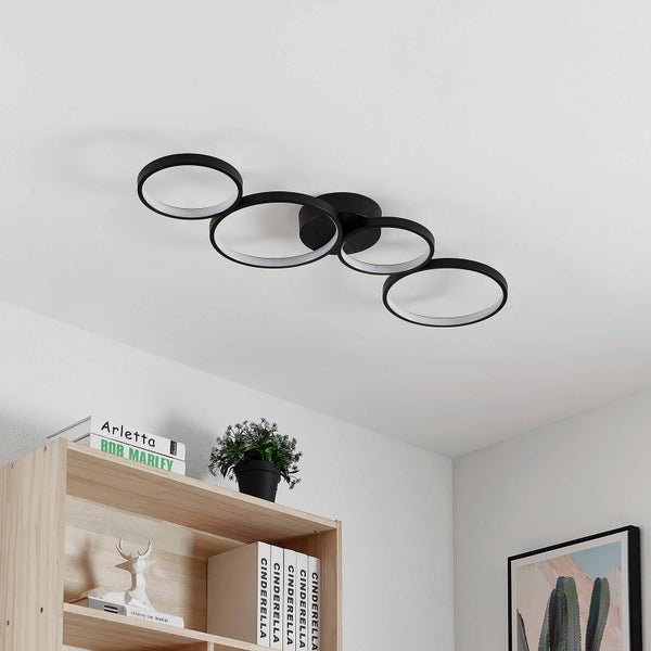Moderne LED-Deckenleuchte mit vier ineinandergreifenden schwarzen Ringen über einem Holzregal in einem hellen, minimalistischen Wohnraum.