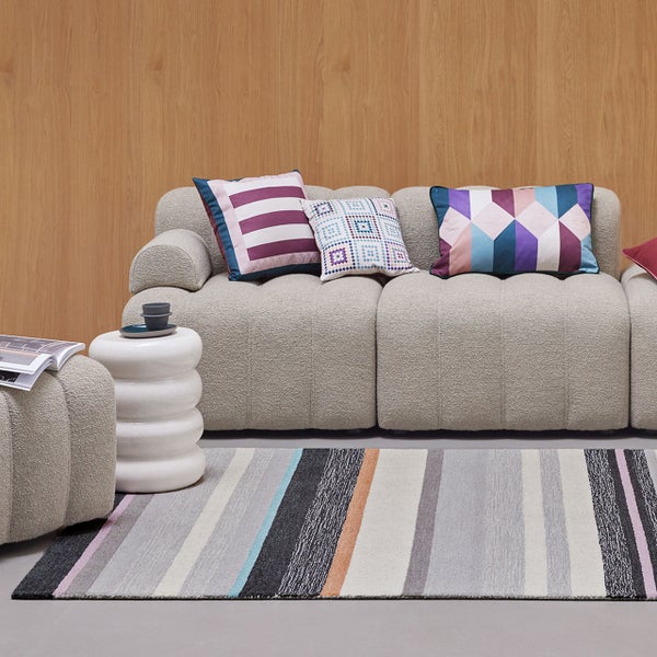 Modernes Wohnzimmer mit hellgrauem Polstersofa, bunten Kissen, gestreiftem Teppich und skulpturalem Beistelltisch vor einer hölzernen Wandpaneele.