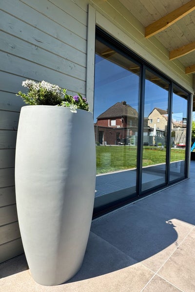 Hoher grauer Pflanzkübel mit Blumen auf einer modernen Terrasse neben einer großen Glasfront, die den Garten widerspiegelt.