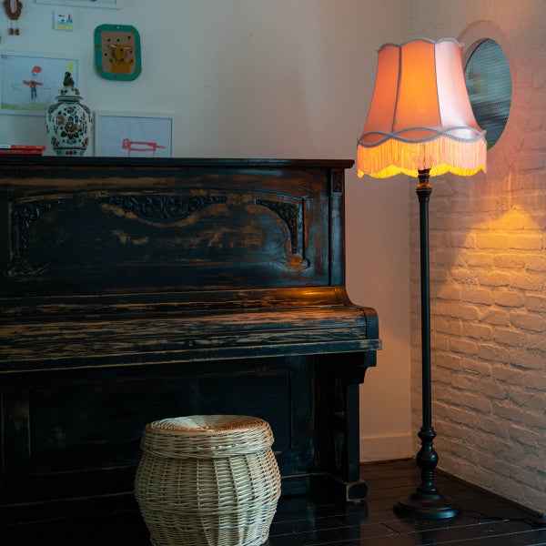 Beleuchtete Stehleuchte mit Fransen-Schirm neben einem dunklen Klavier aus Holz und einem Weidenkorb vor einer Backsteinwand.