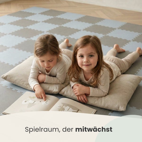 Zwei Kinder liegen auf Kissen auf einer grau-hellgrau karierten Puzzlematte im Spielzimmer und lesen ein Buch.