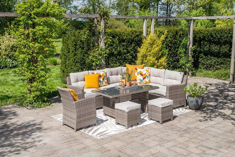Helles Lounge-Set aus Polyrattan mit Ecksofa, Sessel, Tisch und Hockern auf einer Terrasse im Garten mit grüner Hecke und Bäumen.
