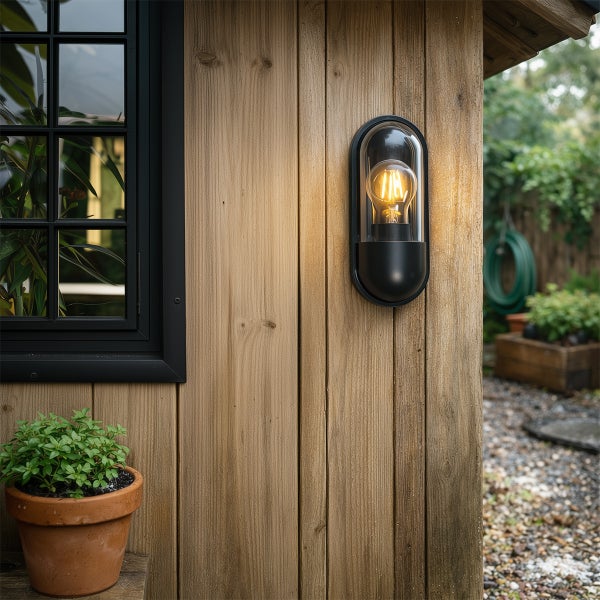 Ovale Außenwandleuchte mit Filamentlampe montiert an einer hölzernen Gartenhauswand neben einem Fenster.