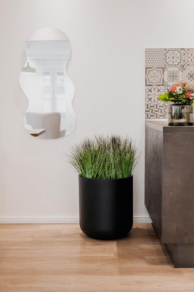 Moderner Wohnraum mit schwarzem zylindrischem Pflanztopf inklusive Kunstgras, wellenförmigem Wandspiegel und grauem Sideboard auf Holzboden.