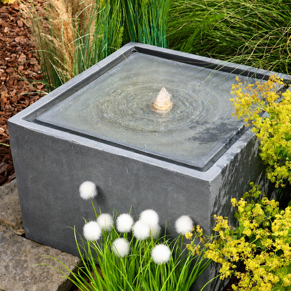 Quadratischer Gartenbrunnen in Steinoptik mit zentralem Wasserspiel inmitten einer bepflanzten Gartenanlage.