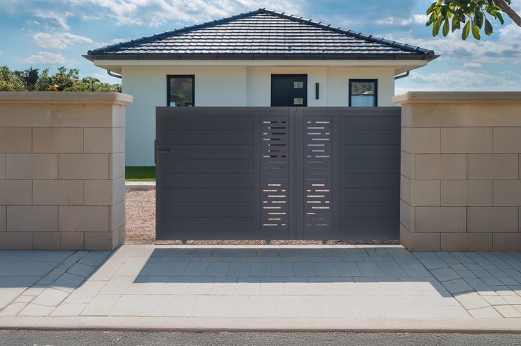 Modernes zweiflügeliges Einfahrtstor in Anthrazit mit horizontalen Lamellen und Dekorausschnitten zwischen hellen Steinpfosten vor einem Wohnhaus.