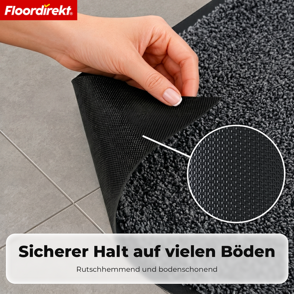 Floordirekt Logo. Eine Hand hebt die Ecke einer Matte auf Fliesen an, um die rutschhemmende Rückseite zu zeigen. Sicherer Halt auf vielen Böden.