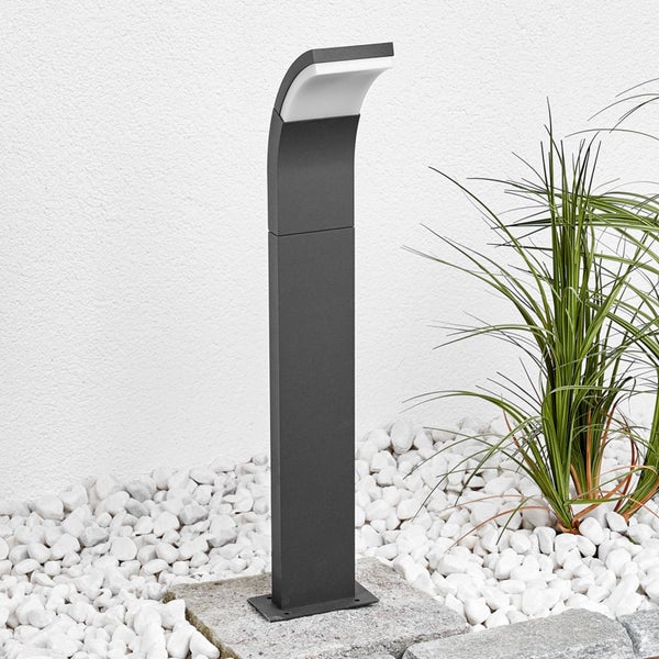 Moderne Wegeleuchte in Anthrazit mit gebogenem Kopf, platziert in einem Gartenbeet mit weißen Steinen und Ziergras.