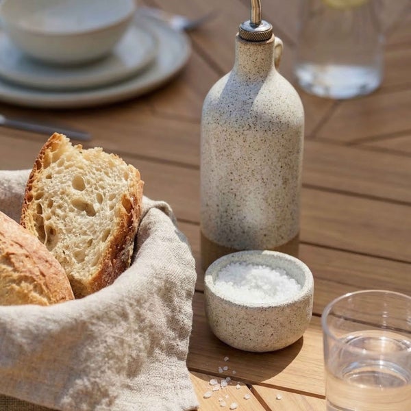 Gedeckter Holztisch mit Brotkorb aus Leinen, Keramikflasche für Öl und kleiner Salzschale mit grobem Meersalz.