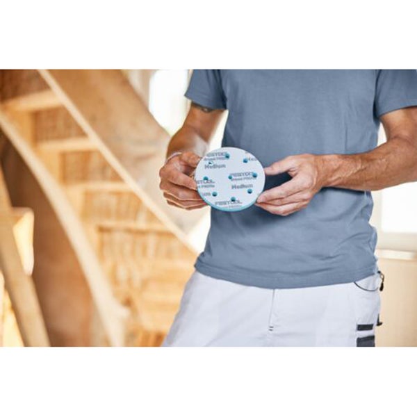 Person hält einen runden Festool Schleifteller Medium mit Multi-Jetstream-Lochung vor einer Treppe aus hellem Holz.