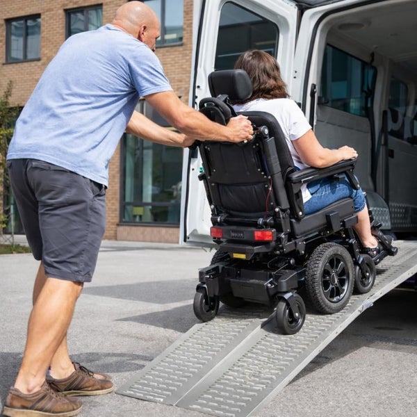 Ein Mann schiebt eine Frau in einem elektrischen Rollstuhl über eine Auffahrrampe in einen Transporter.