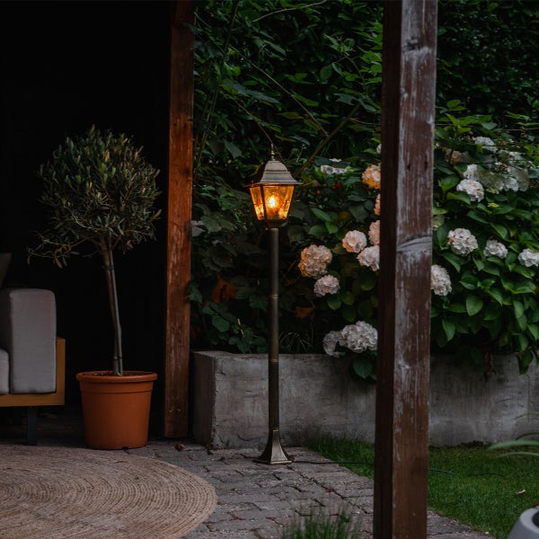 Außen-Sockelleuchte im Laternen-Design mit warmem Licht auf gepflasterter Terrasse neben einem Olivenbaum und weißen Hortensien im Garten.