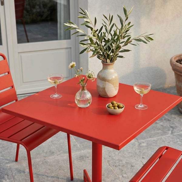 Quadratischer roter Gartentisch aus Metall mit passenden Stühlen auf einer hellen Terrasse, dekoriert mit Vasen und Olivenzweigen.