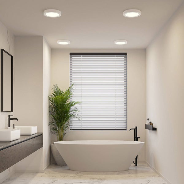 Modernes, helles Badezimmer mit freistehender Badewanne, weißen Jalousien am Fenster, einer großen Zimmerpflanze und Einbauleuchten an der Decke.