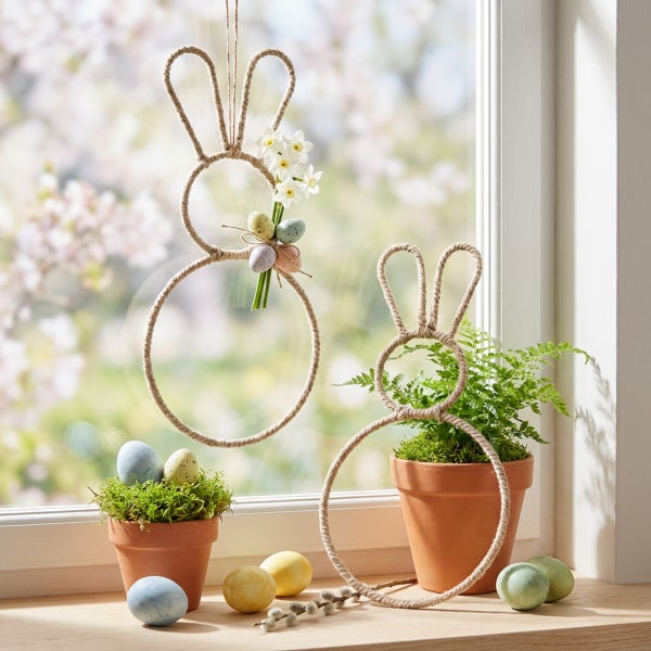 Osterdekoration mit zwei hängenden und stehenden Hasen aus Sisalseil mit Ostereiern und Blumen auf einem Fensterbrett