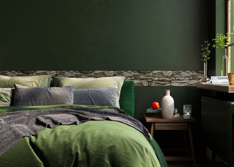 Schlafzimmer mit dunkelgrüner Wand und dekorativer Naturstein-Bordüre, Bett mit grüner Bettwäsche neben einem Holznachtisch mit Dekoration.