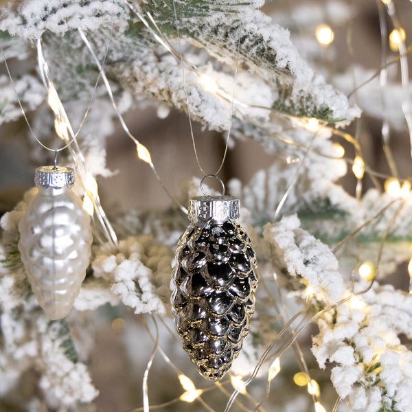 Zwei tannenzapfenfoermige Christbaumkugeln haengen an einem verschneiten Weihnachtsbaum mit einer Lichterkette.