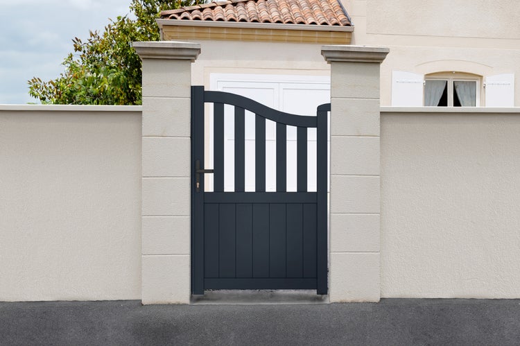Dunkelgraues Gartentor mit geschwungener Oberkante und vertikalen Stäben zwischen zwei hellen Steinpfosten vor einer hellen Mauer.