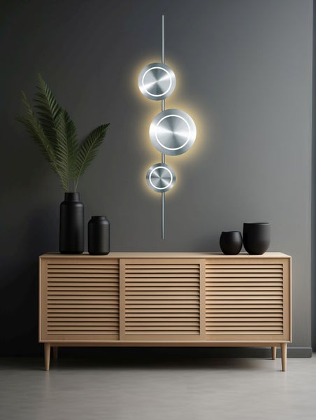 Moderne LED Wandleuchte mit drei runden Metallelementen an einer vertikalen Stange über einem Sideboard aus hellem Holz.