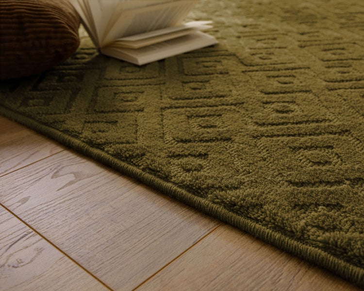 Grüner Teppich mit geometrischem Rautenmuster in einem hellen Wohnzimmer vor einem beigen Sofa und Holzmöbeln.