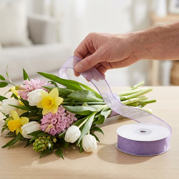 Hand wickelt ein lila Schmuckband um einen frühlingshaften Blumenstrauß mit Narzissen und Hyazinthen auf einem Holztisch.