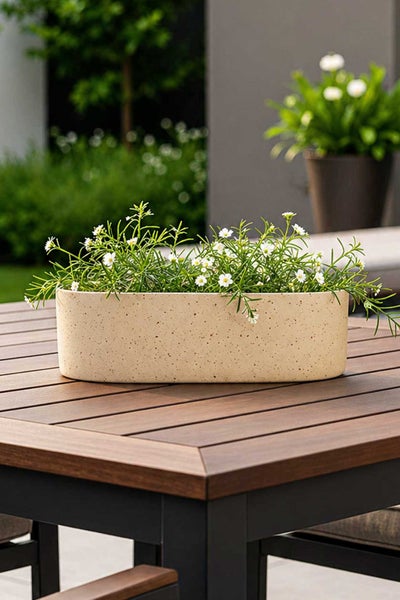 Ovaler Pflanzkasten mit gesprenkelter Oberfläche und Blumen auf einem Gartentisch aus Holz.