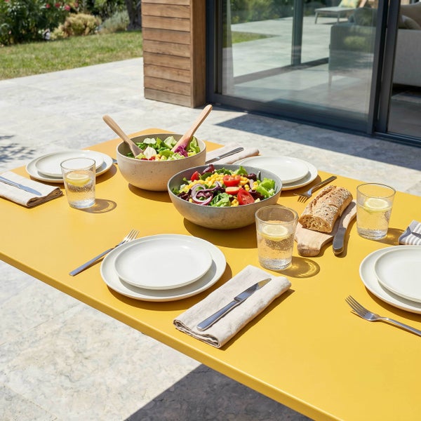 Gedeckter gelber Gartentisch mit Salatschüsseln, Tellern, Gläsern und Besteck auf einer Terrasse.