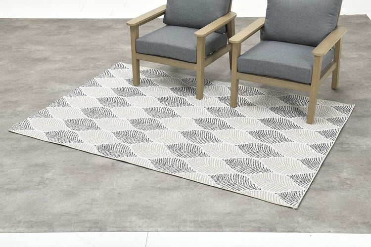 Flachgewebter Outdoor-Teppich mit Blattmuster unter zwei Gartenstühlen aus Holz mit grauen Polstern.